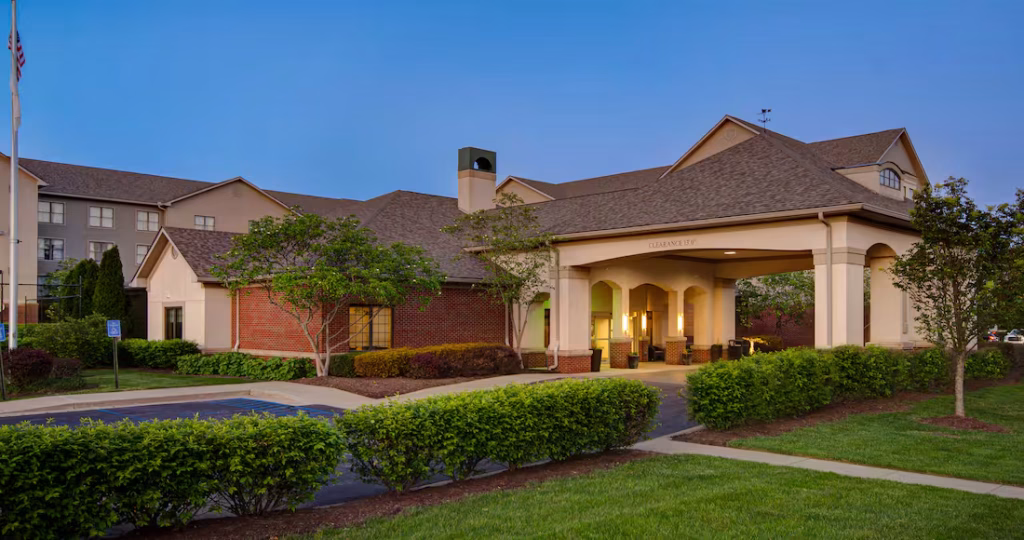 Exterior view of a hotel building at dusk, featuring a covered driveway entrance with warm lighting, landscaped shrubs, and trees in the foreground.