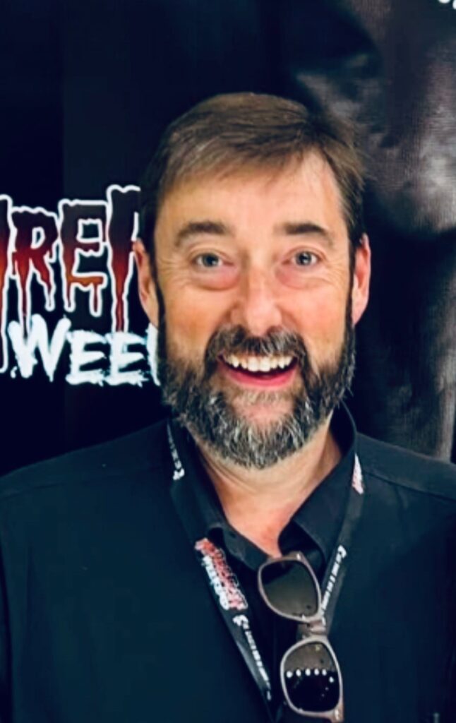 Man with short brown hair and beard smiling in front of a ScareFest Weekend banner, wearing a black shirt, event lanyard, and sunglasses hanging from his collar.