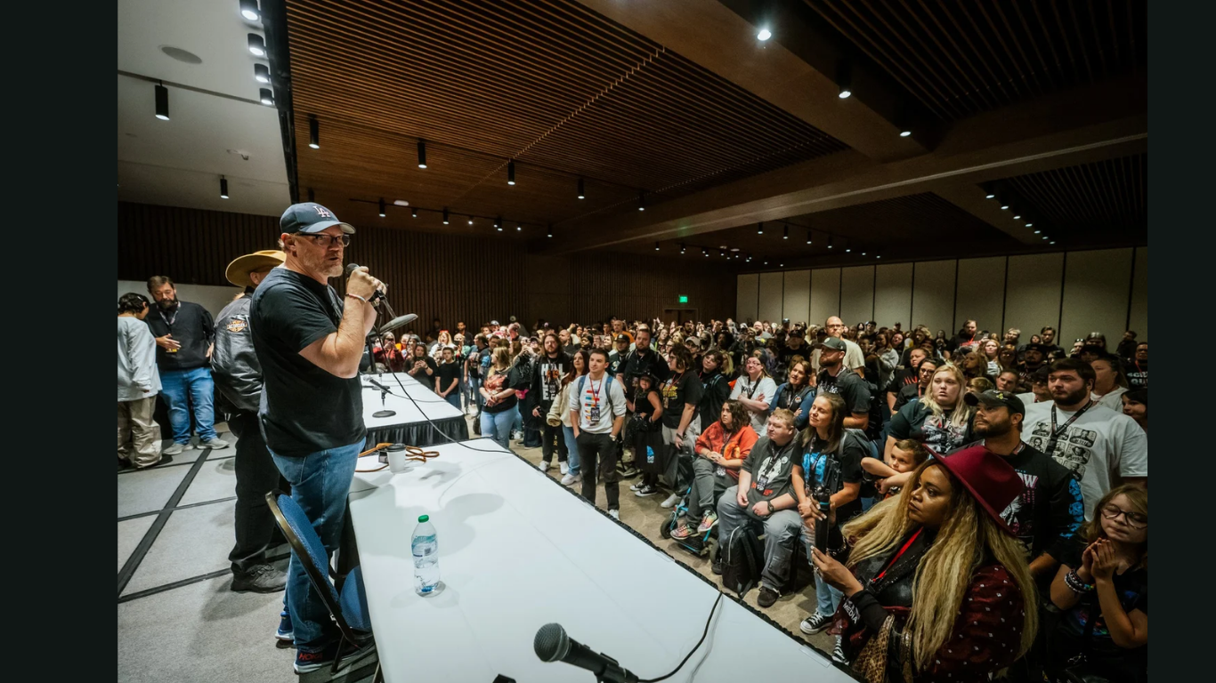 Actor Matthew Lillard speaks into a microphone onstage during a packed convention panel. He stands at a long table with water bottles and microphones, while a large audience fills the room, listening and watching intently.