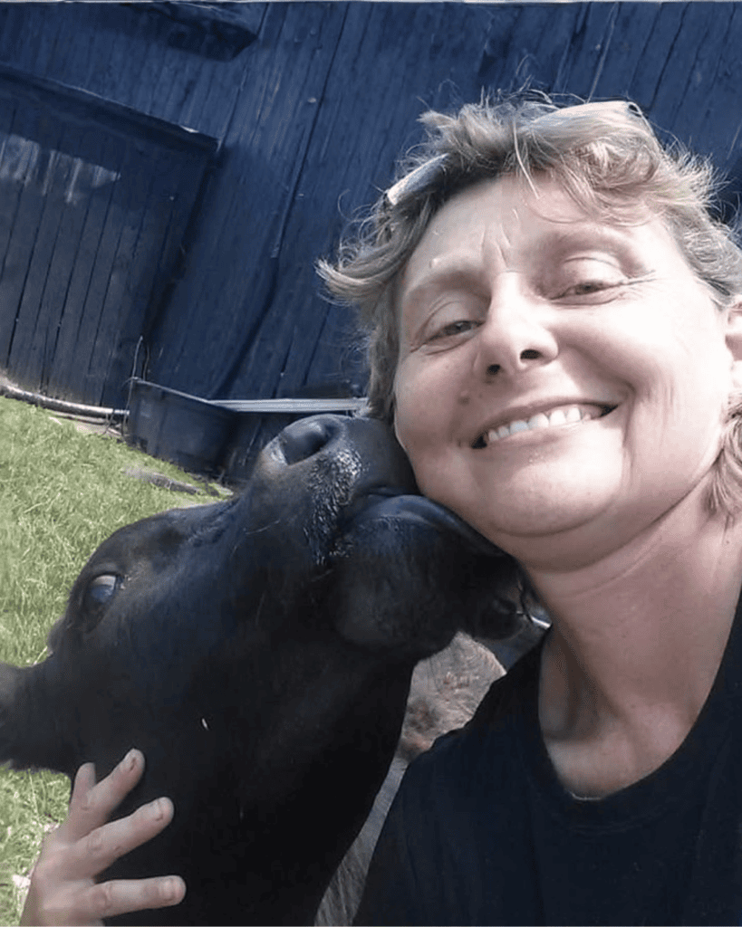 Anita: Anita smiling outdoors as a black cow affectionately nuzzles her cheek.