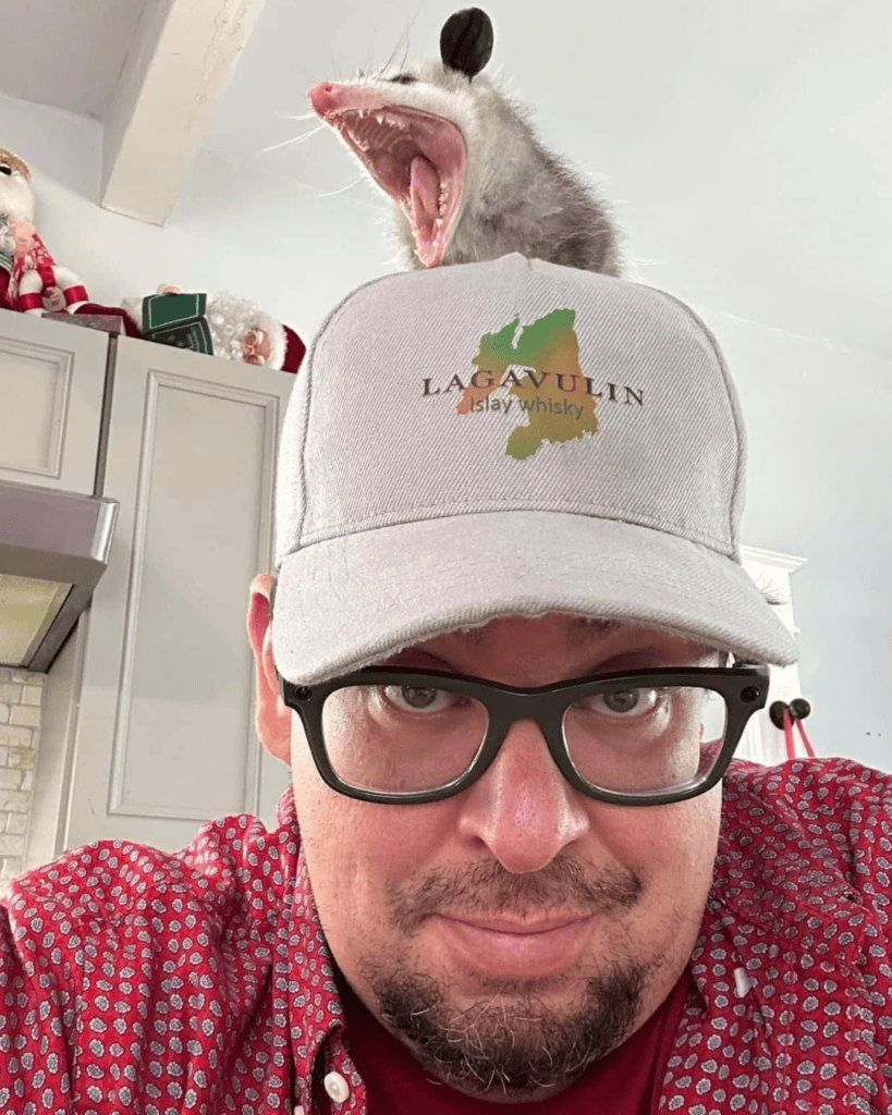 Brandon: Brandon wearing glasses and a gray baseball cap, with a wide-mouthed opossum perched behind him.
