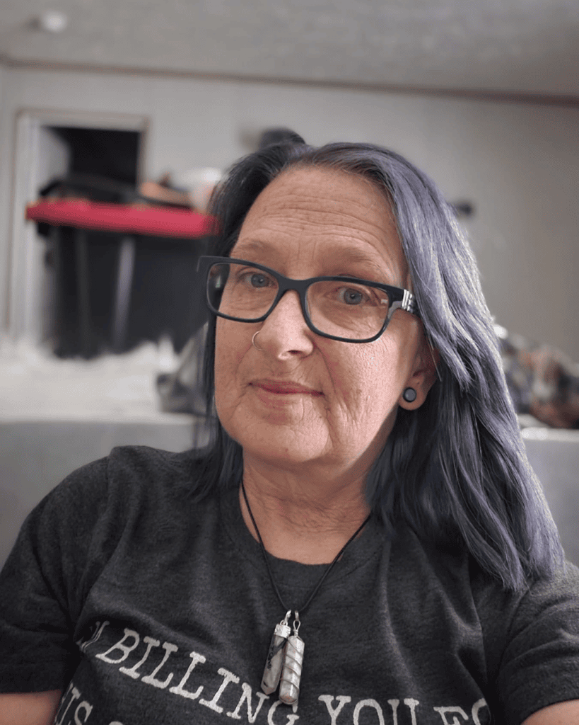 Portrait of Gail wearing glasses with long blue-purple hair, a nose ring and crystal pendant necklace.
