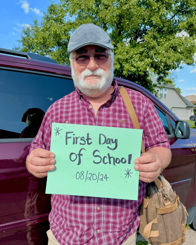 Joe wearing a flat cap and plaid shirt, standing beside a vehicle and holding a “First Day of School” sign.