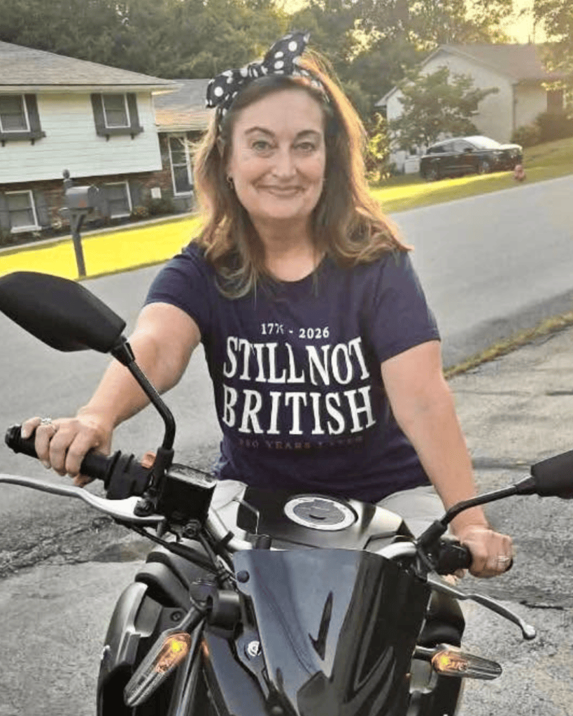 Lori smiling while seated on a motorcycle, wearing a polka-dot bow and “Still Not British” T-shirt.