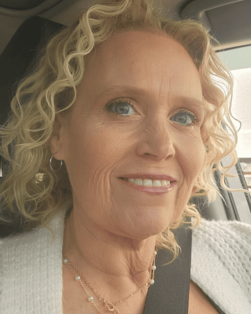 Close-up portrait of Megan smiling in a vehicle with curly blonde hair and layered necklaces.