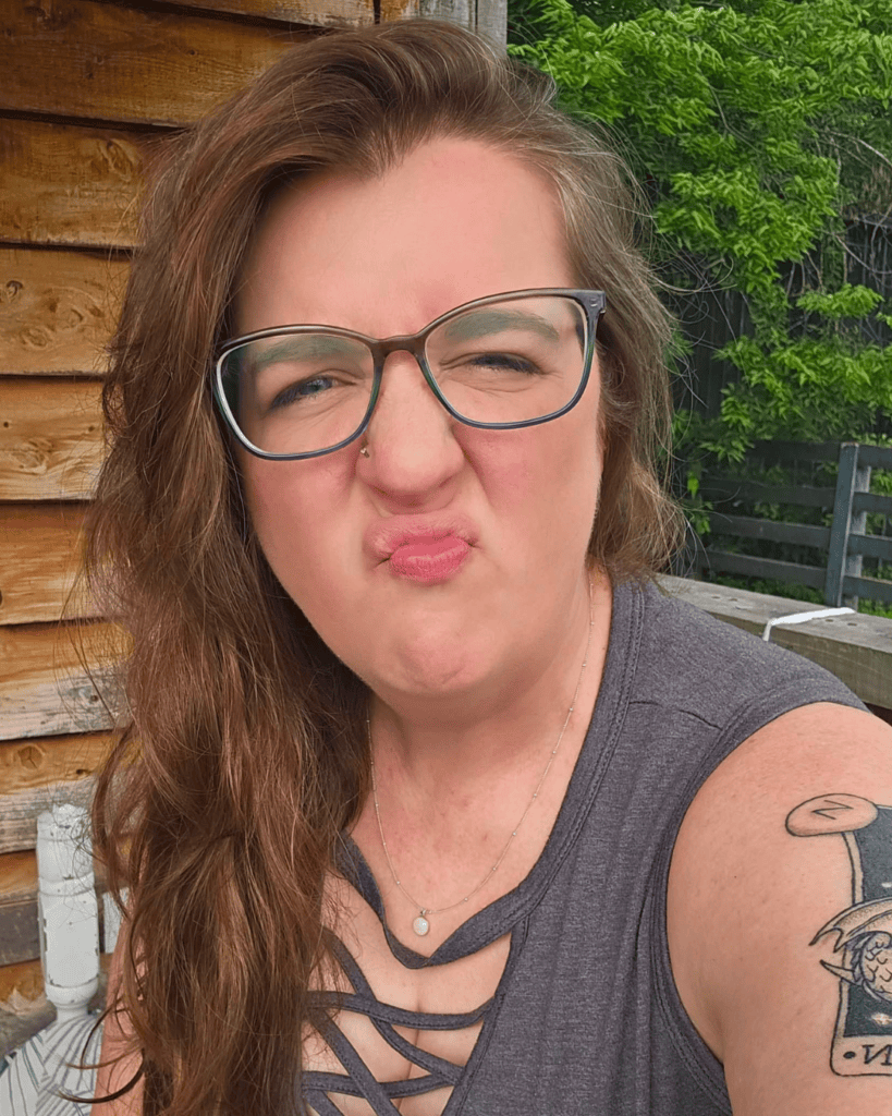 Meme wearing glasses and making a playful face outdoors, with long brown hair and an upper-arm tattoo.