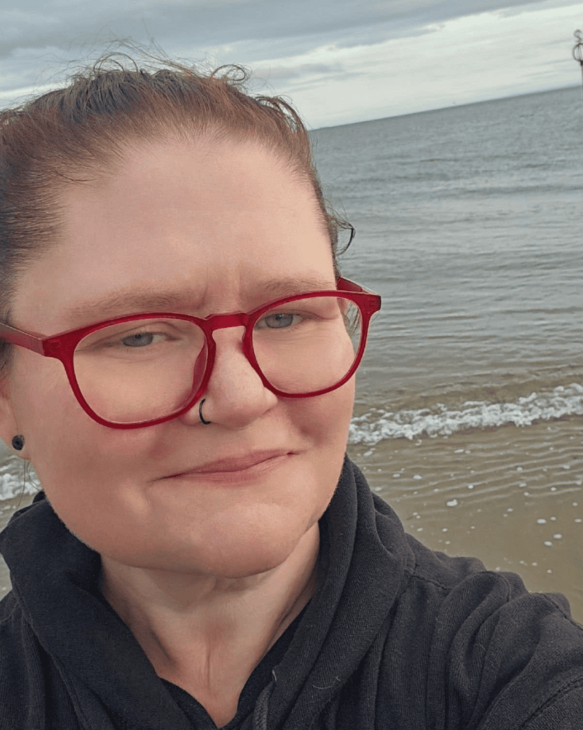 Merrideth wearing red glasses and a nose ring, taking a selfie beside the ocean.