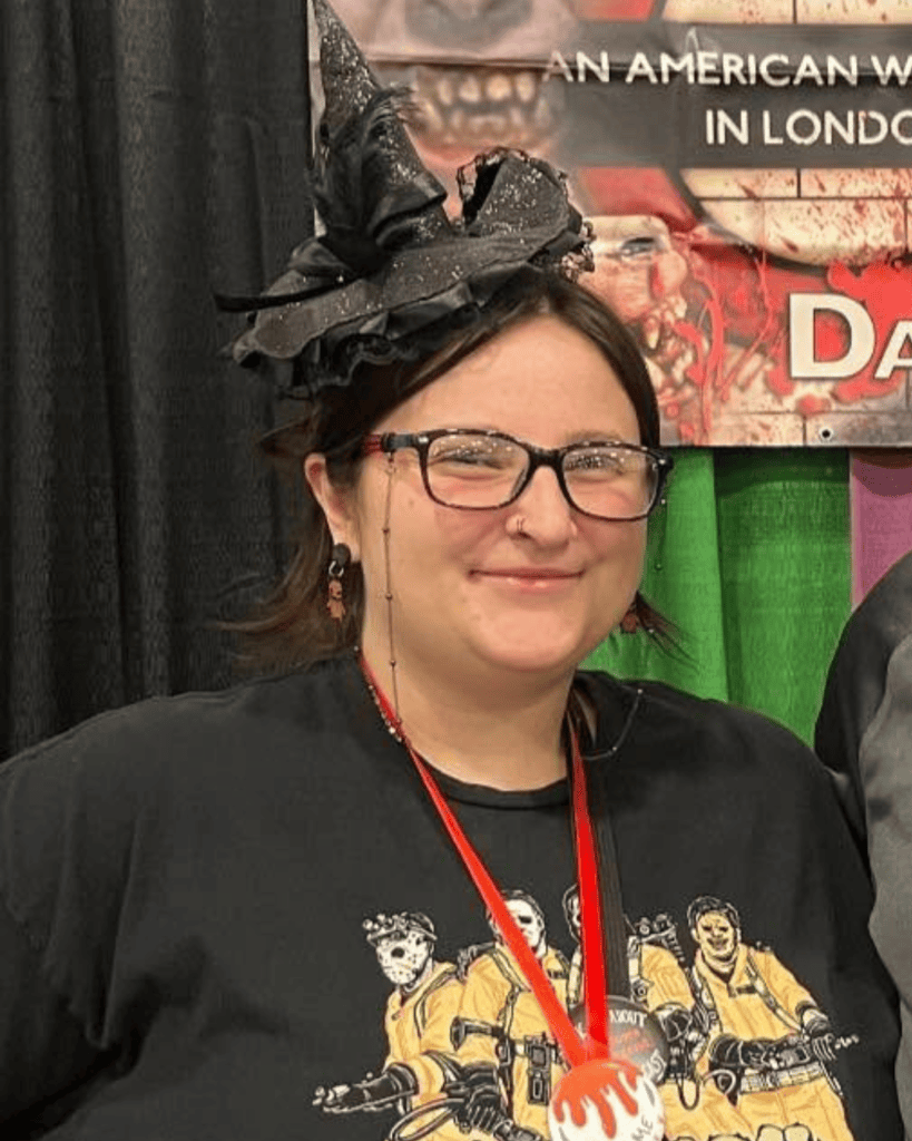 Samantha smiling in glasses and a miniature witch hat, wearing a horror-themed T-shirt and red lanyard.